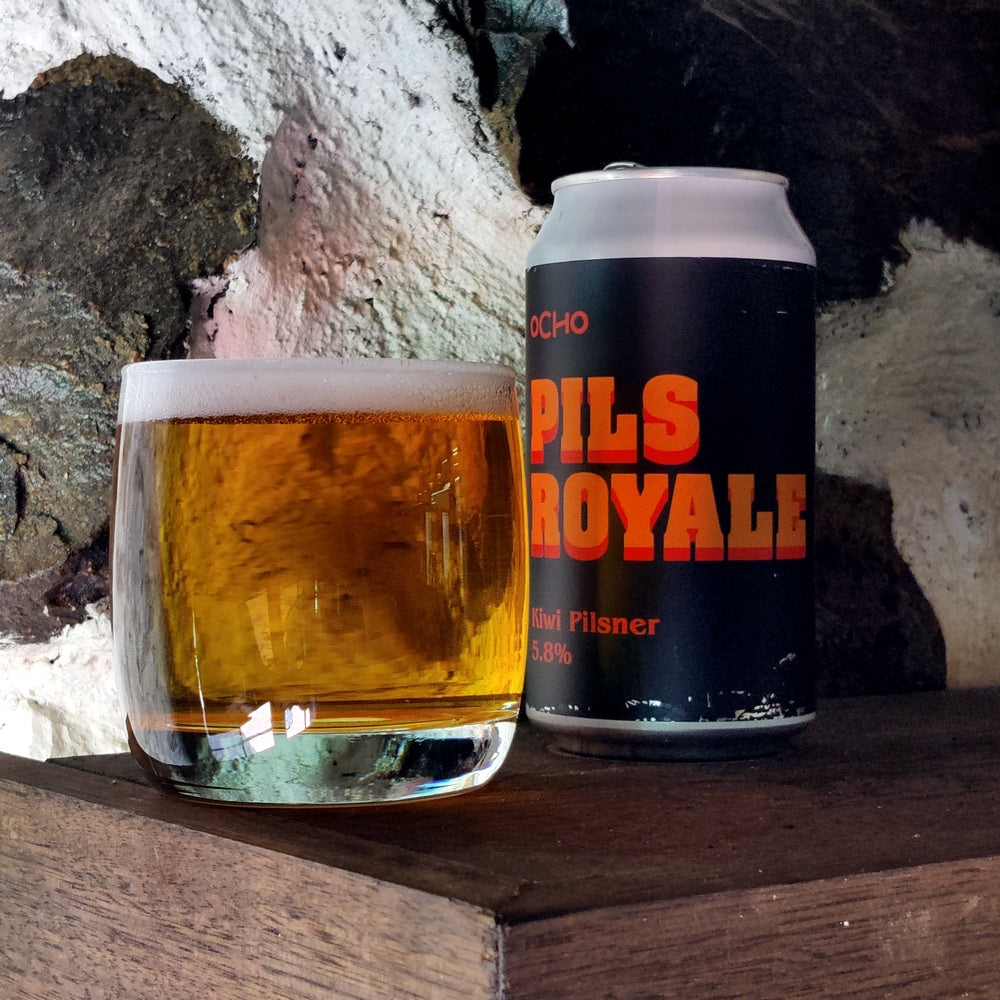 This is a photo of a can of Ocho Pills Royale with a glass of beer next to it that has already been poured. This Pilsner is set infront of some stone work