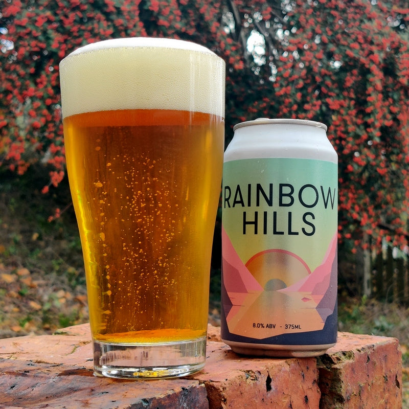 This is a photo of a can of rainbow hills beer and a schooner glass on a retaining wall in the garden. The Schooner glass of beer is full of the Double IPA rainbow hills.